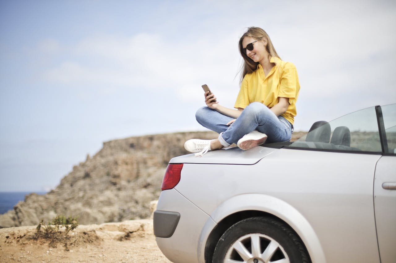Smiling young woman sitting on a car at the beach in Mgarr, Malta using a smartphone.